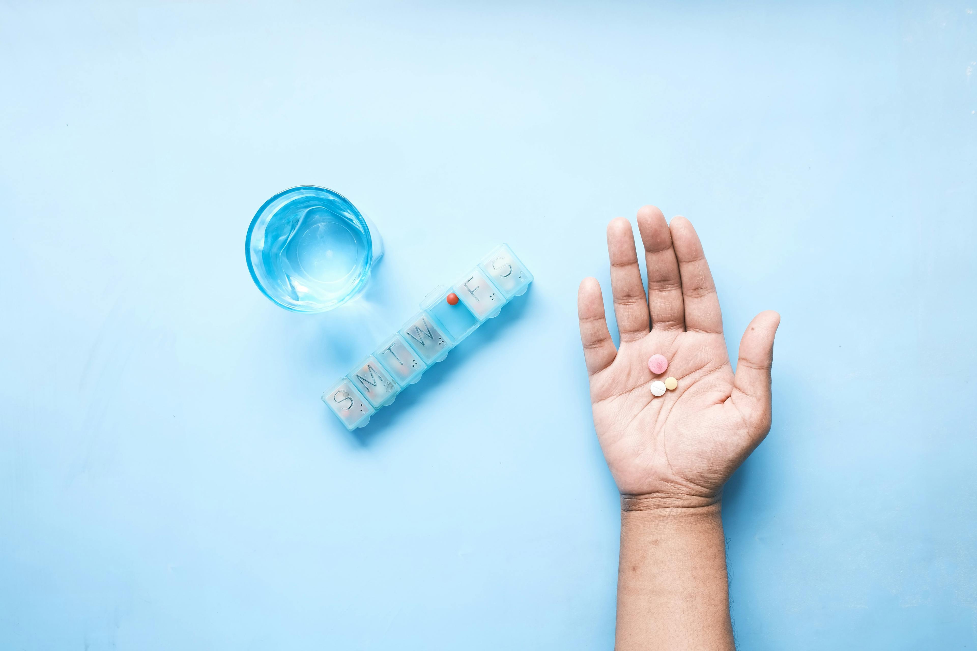 A hand holding three pills, next to a pill container and a glass of water.
