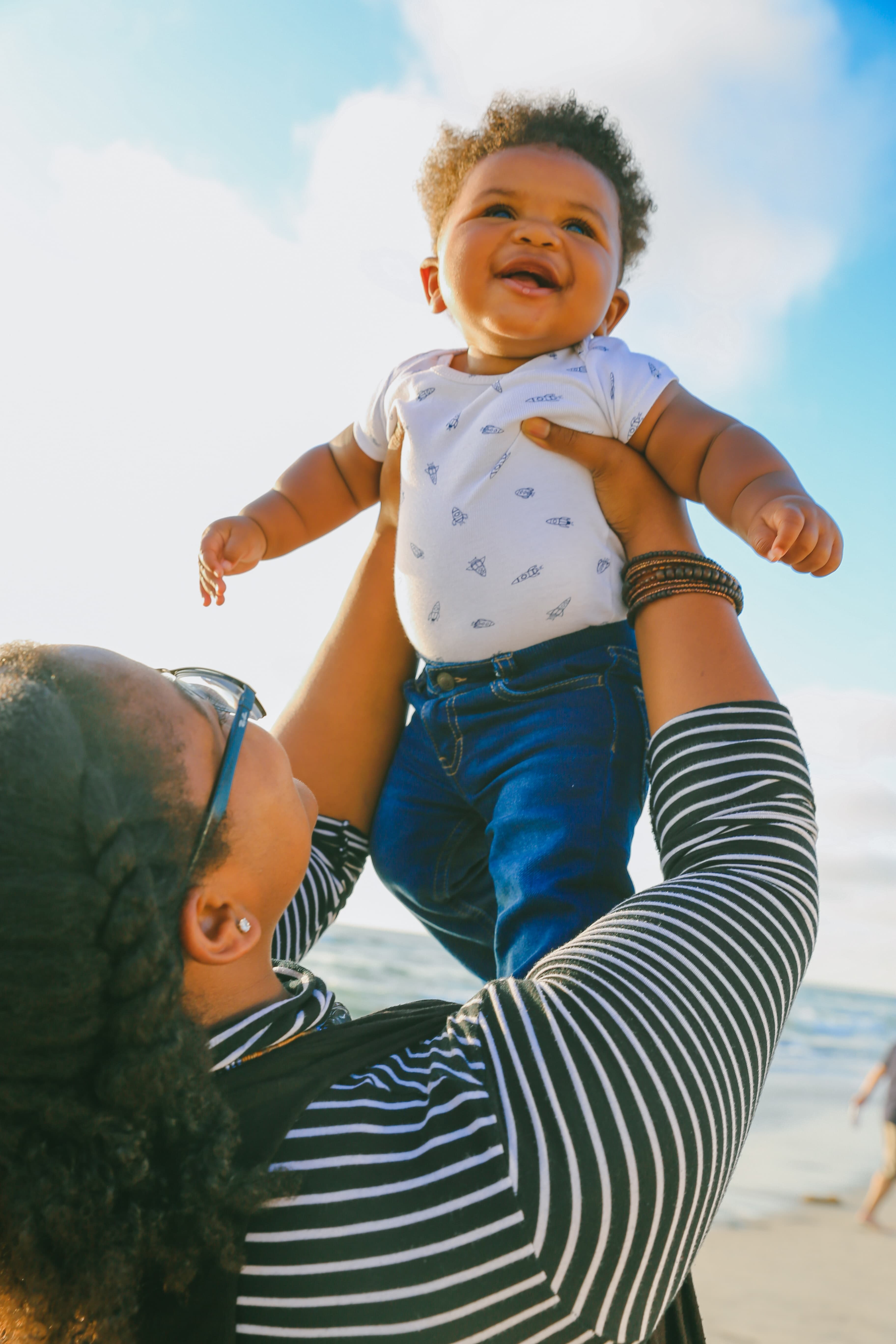 A smiling baby held up to the sky by his mom.