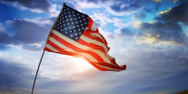 An American flag waves in the air with a sun setting in the background.