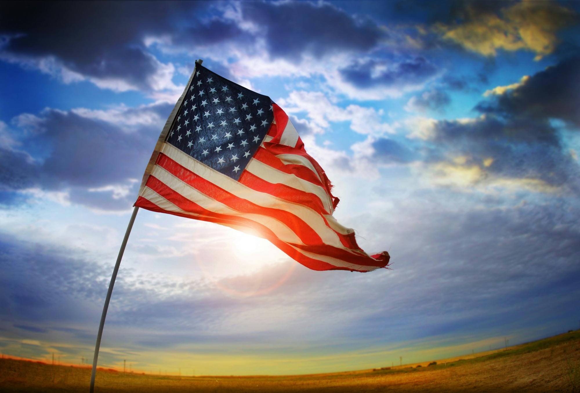 An American flag waves in the air with a sun setting in the background.