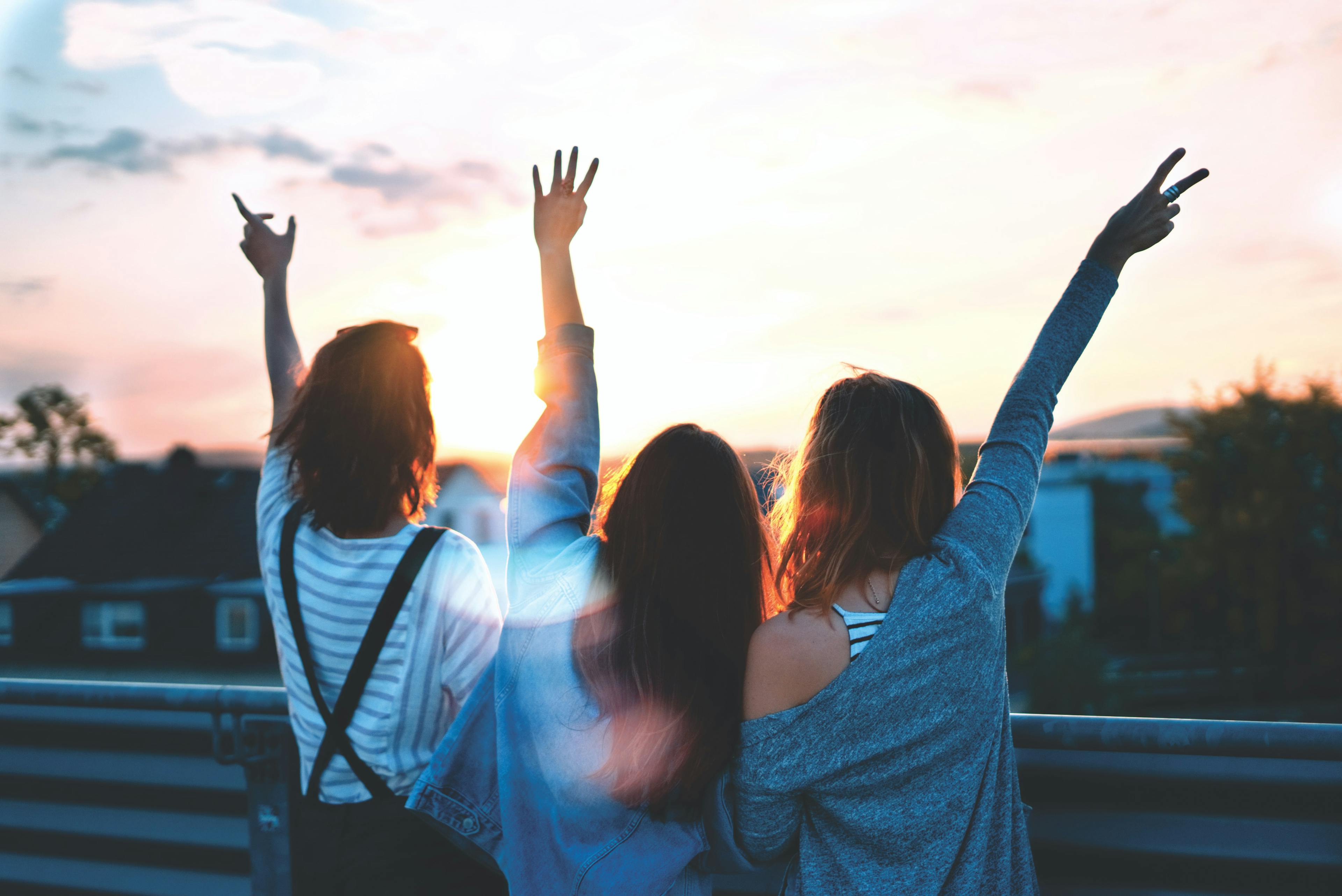 Three friends stand on a roof at sunset, their backs facing the camera. Their arms are outstretched in celebration.