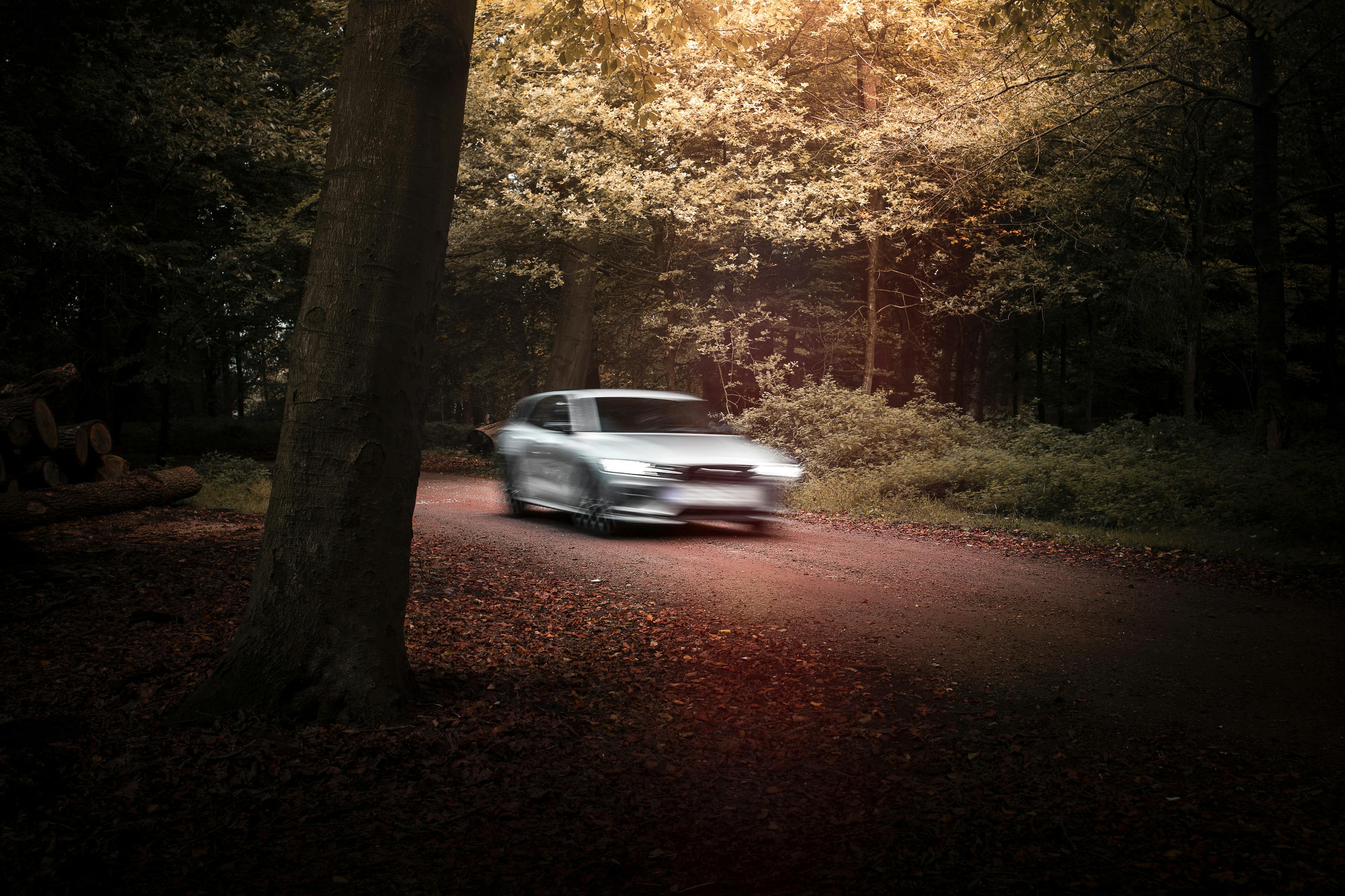 A silver sedan drives on a deserted dirt road surrounded by leafy green trees. The car is blurry and out of focus, while the surrounding forest is clear and in focus.