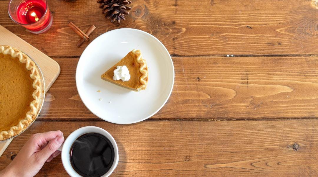 Close-up of a wooden table, on top of which sits pumpkin pie, pinecones, cinnamon sticks, a lit candle, and a hand holding a mug of coffee.
