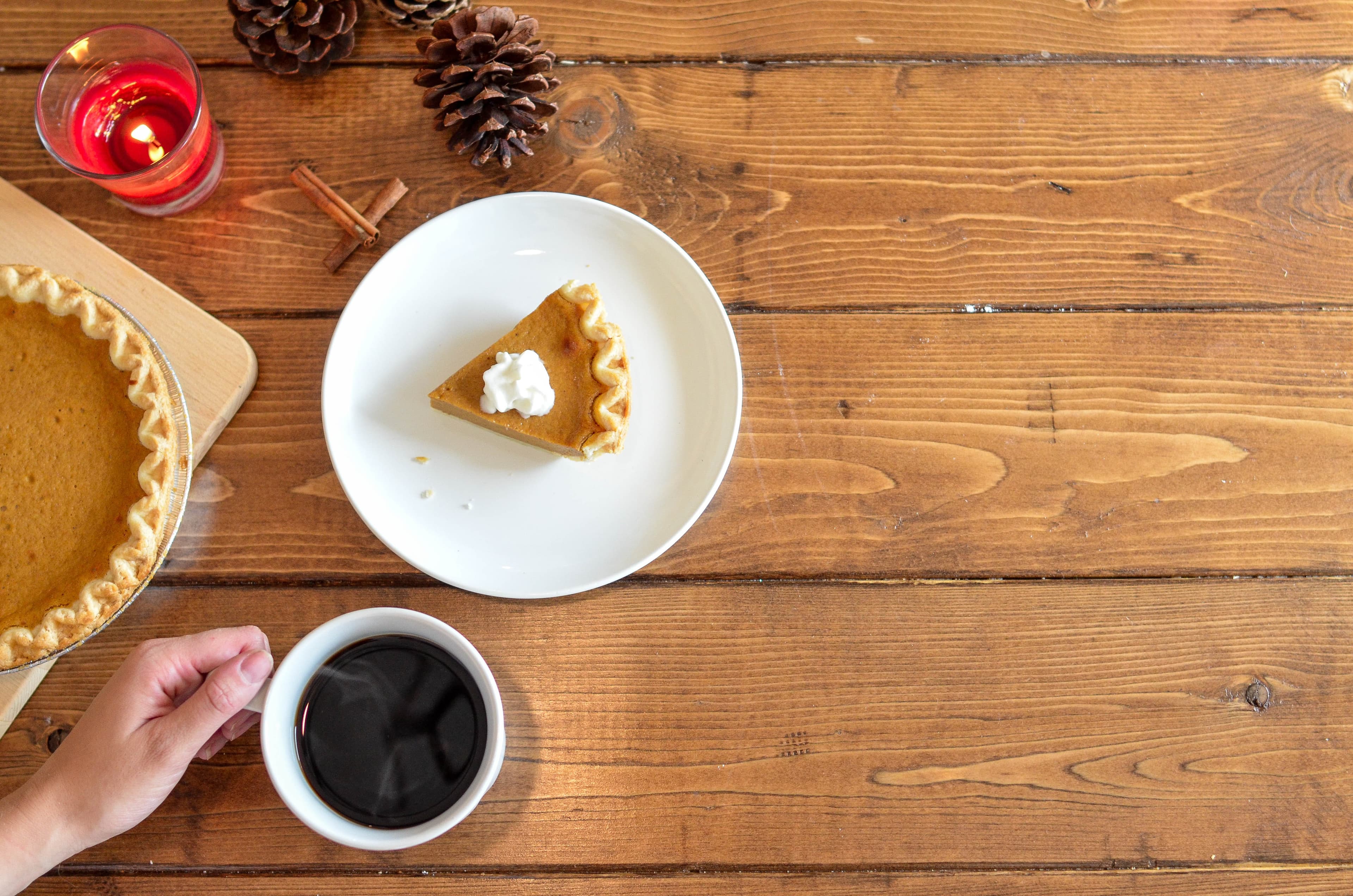 Close-up of a wooden table, on top of which sits pumpkin pie, pinecones, cinnamon sticks, a lit candle, and a hand holding a mug of coffee.