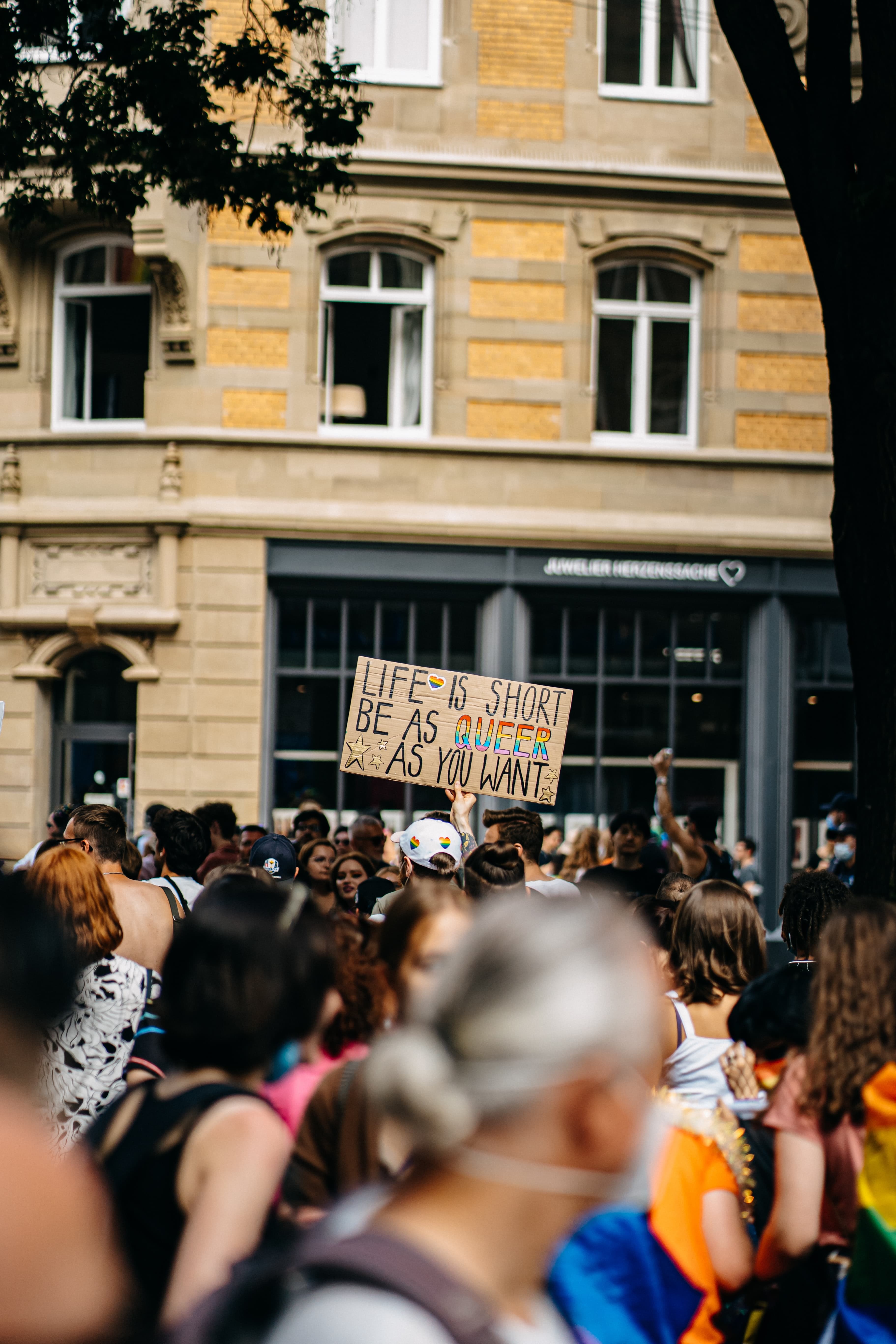 A crowd of blurry people at a pride march in a city. In focus is a cardboard sign with the hand-painted words "Be as queer as you want."