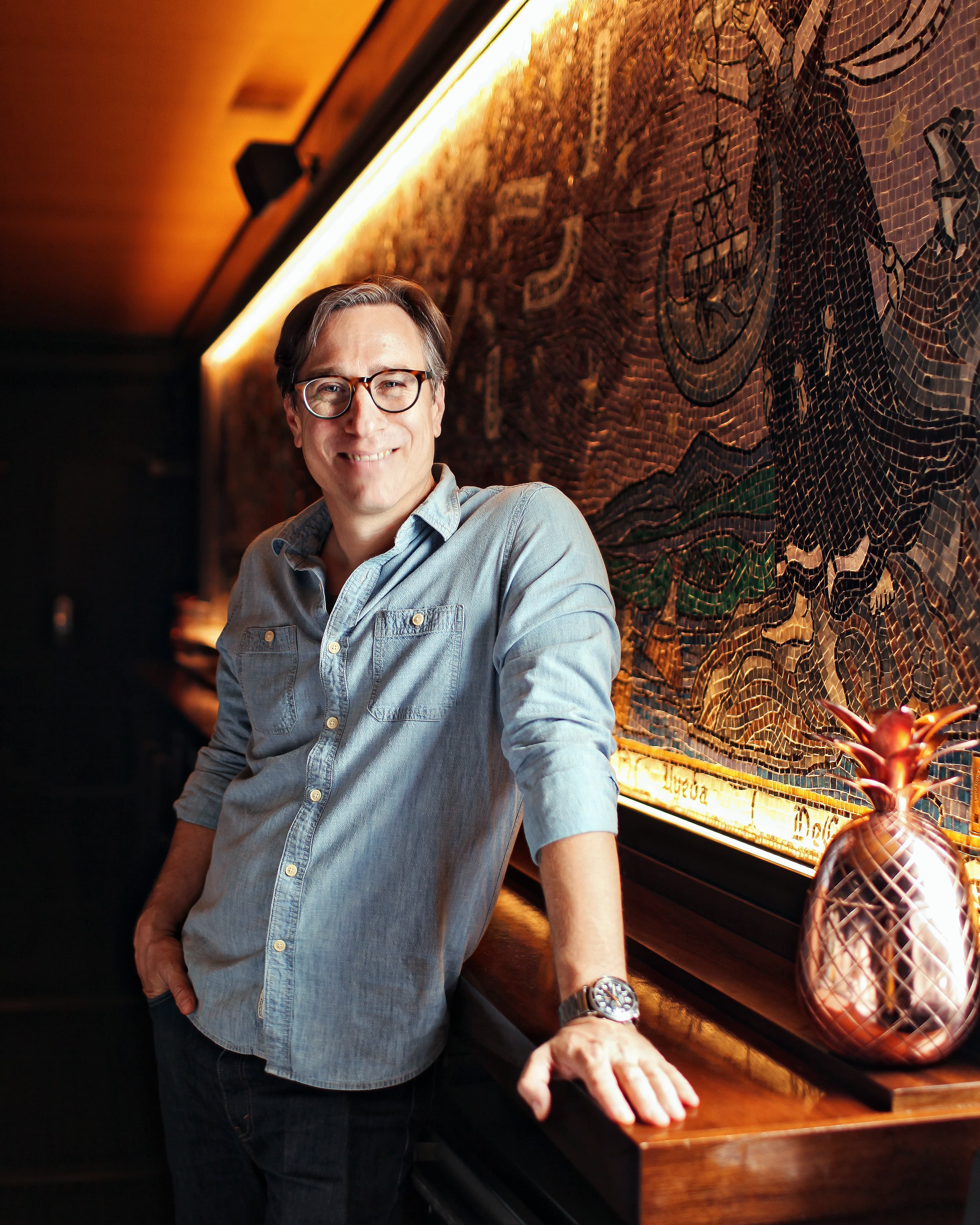 A smiling white man in a light blue button-up shirt and glasses smiles at the camera while leaning against a table.