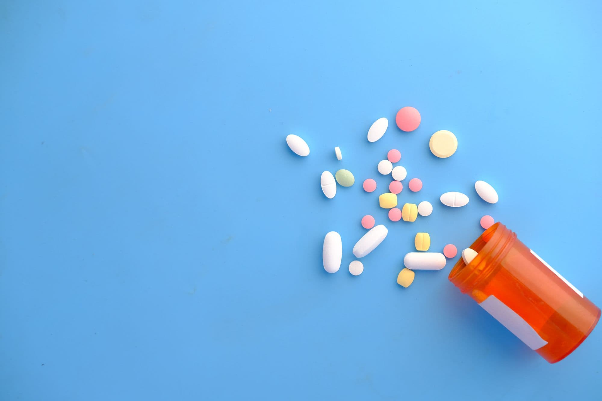 Orange prescription bottle lying on its side with various pills of different sizes and colors spilling out onto a light blue background.