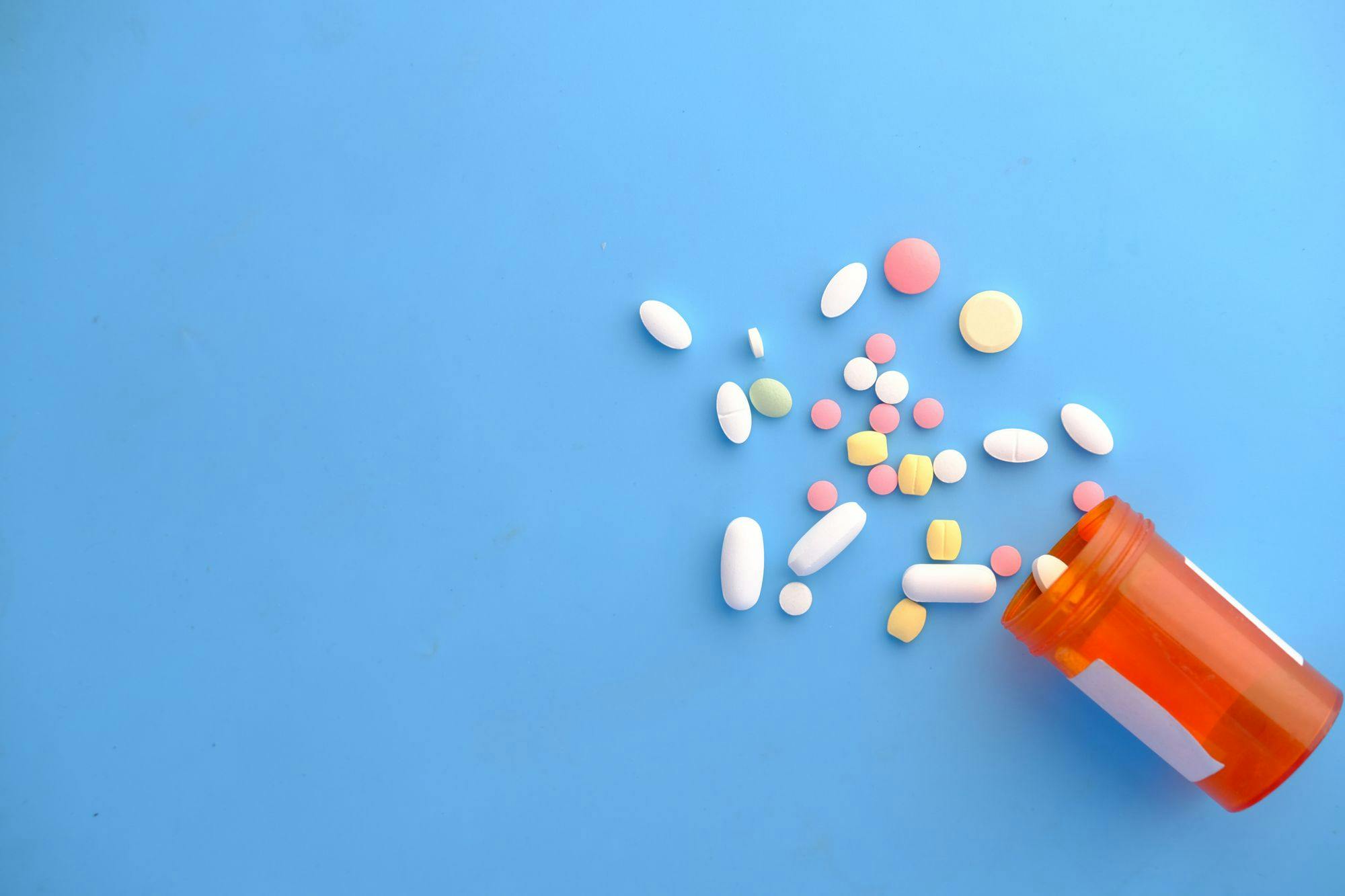 Orange prescription bottle lying on its side with various pills of different sizes and colors spilling out onto a light blue background.