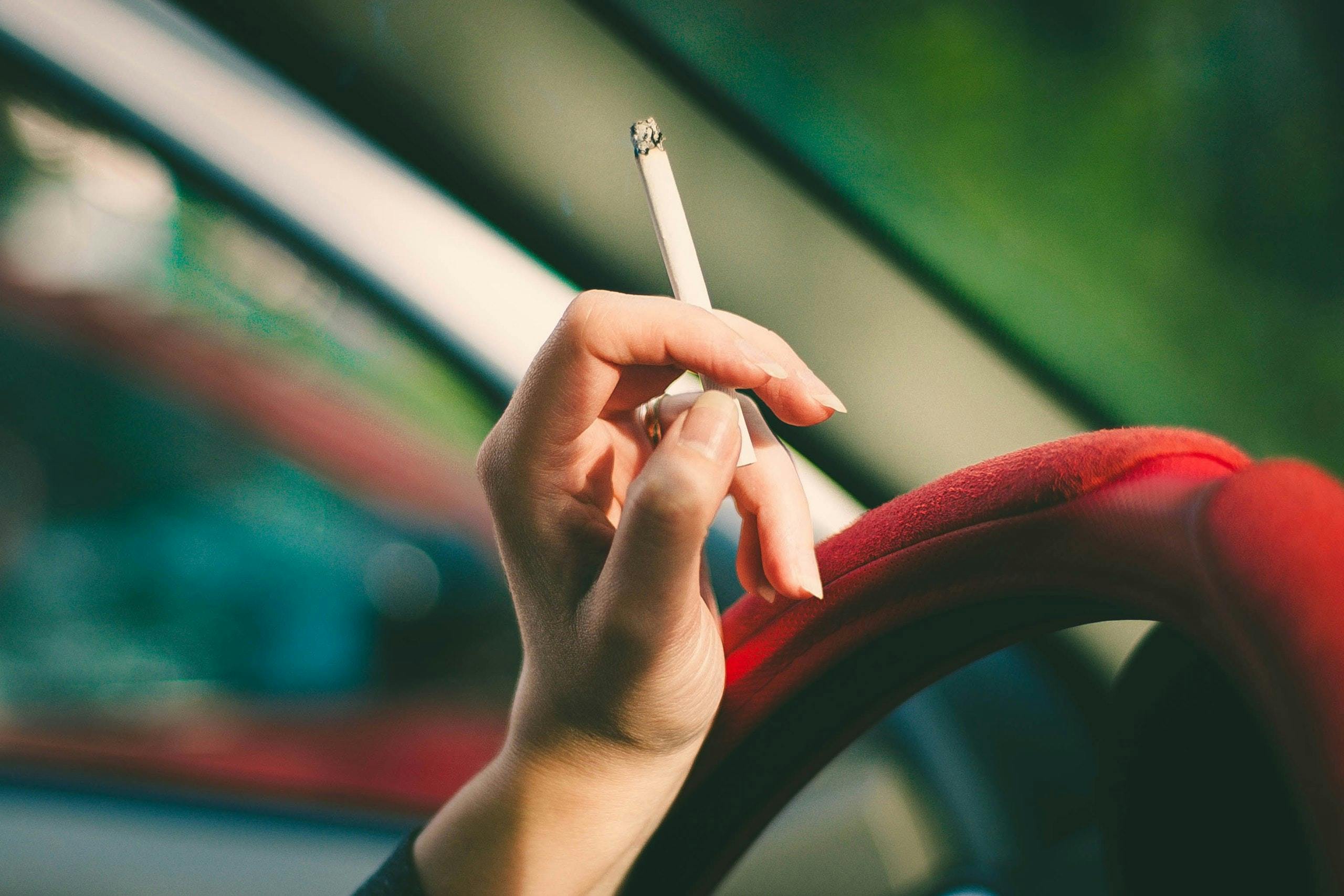 A close-up of a woman's hand resting on a car steering wheel while holding a cigarette.