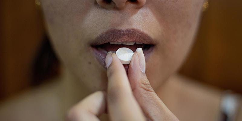 The bottom half of a woman's face is visible. Her hand holds a white pill to her parted mouth.
