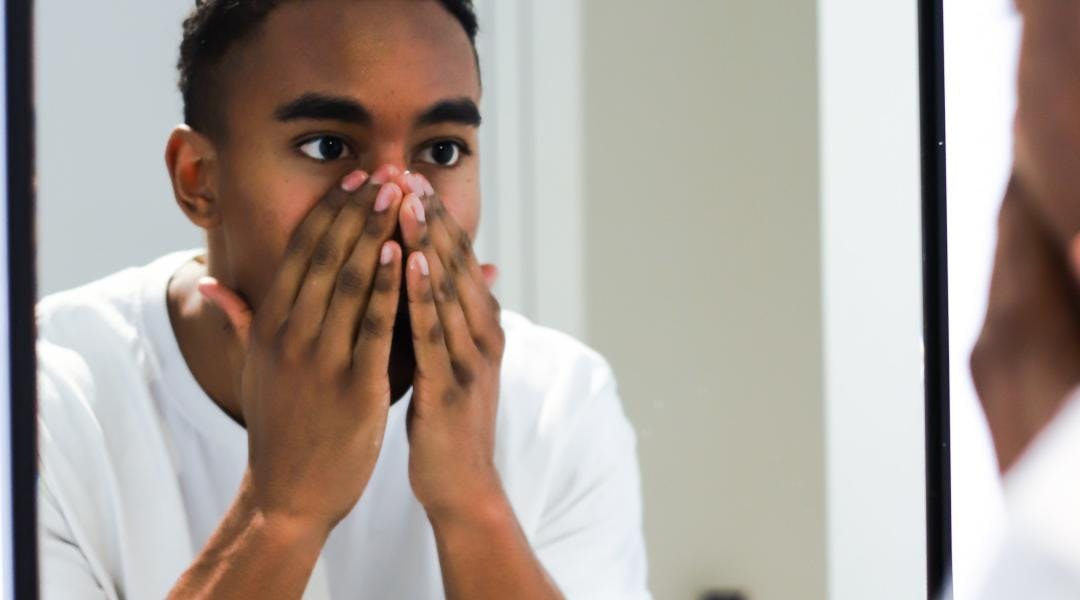 A young Black man looks at his reflection in the mirror. His hands are covering his nose, mouth, and chin.
