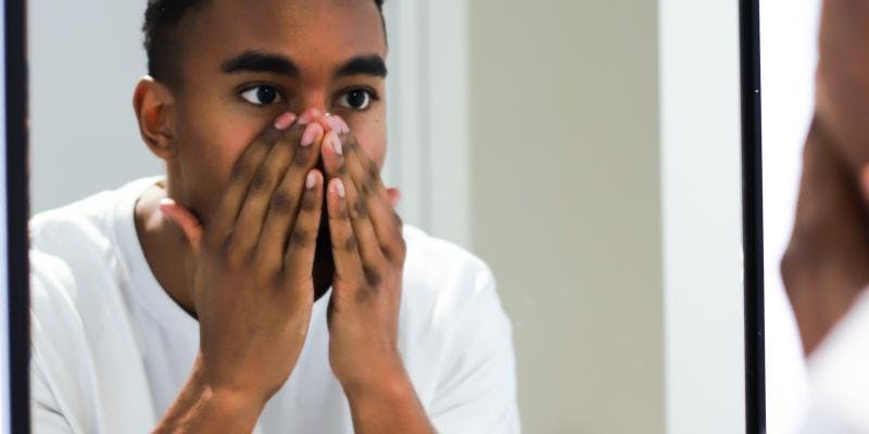 A young Black man looks at his reflection in the mirror. His hands are covering his nose, mouth, and chin.