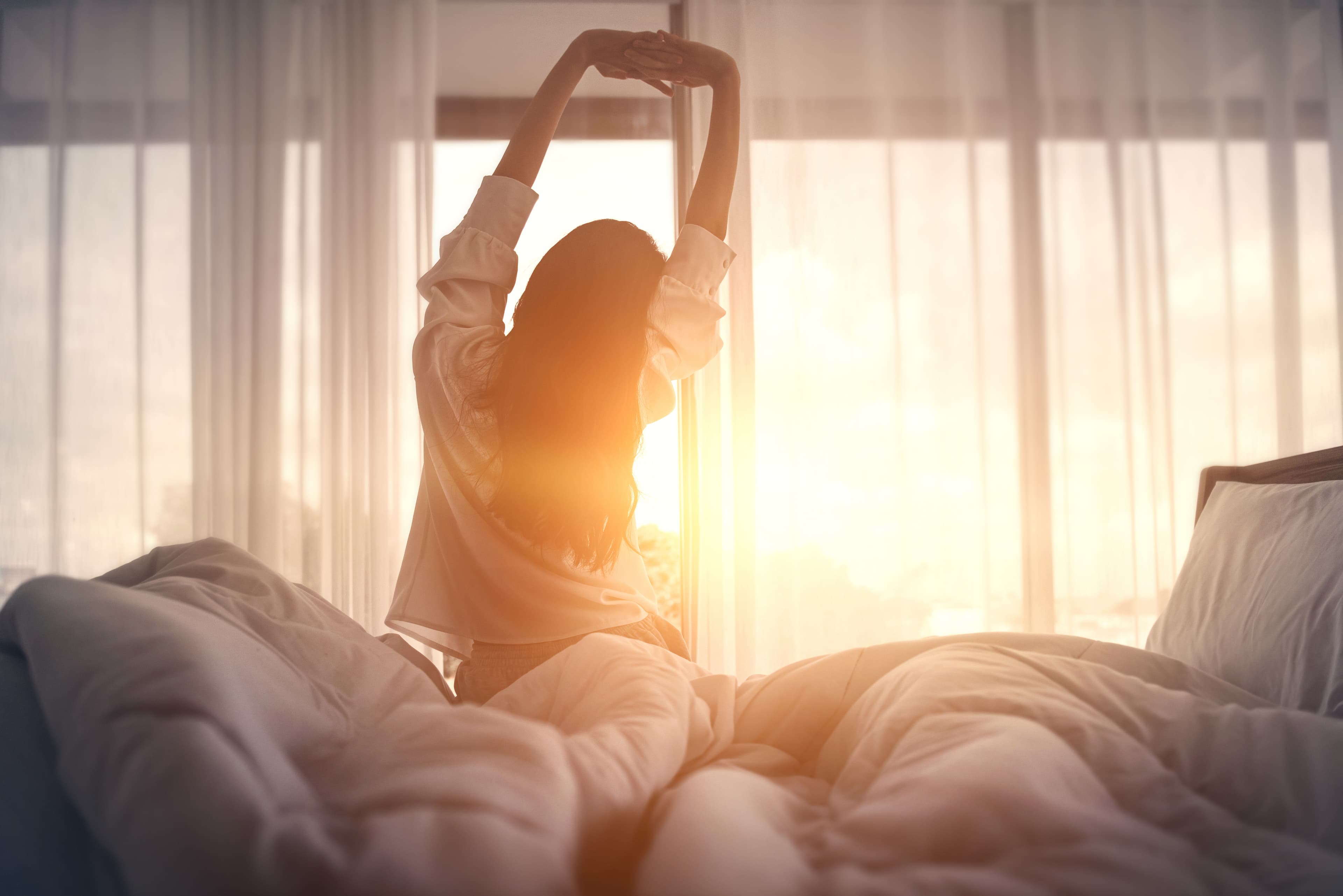 A woman getting out of bed during a sun-drenched dawn and stretching happily.
