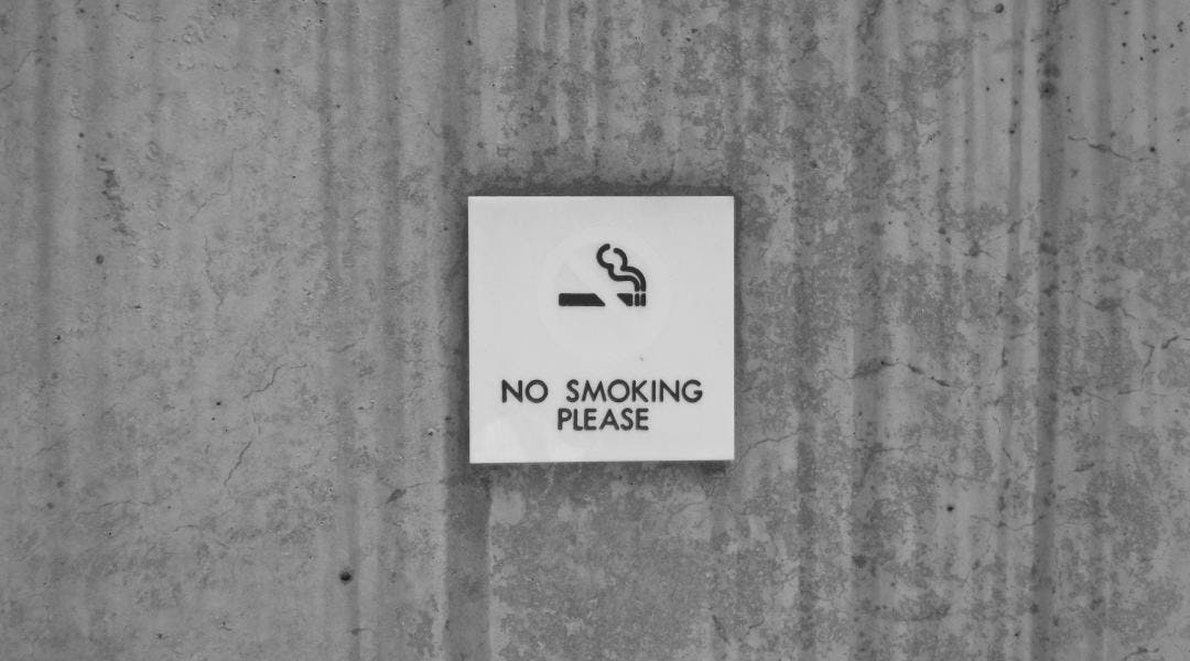 A gray cement wall with a white sign that says "no smoking please" under an illustration of a lit cigarette.