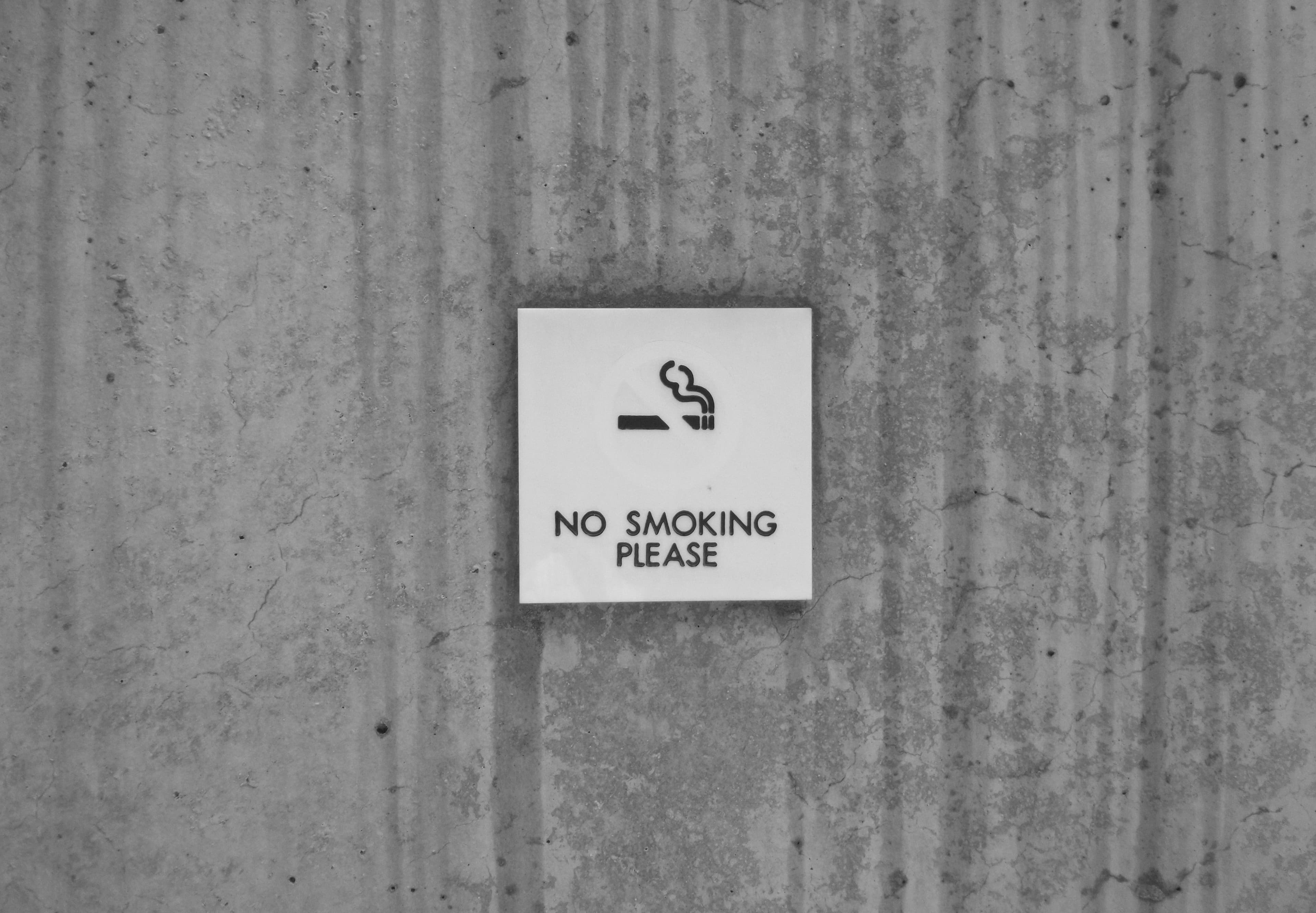 A gray cement wall with a white sign that says "no smoking please" under an illustration of a lit cigarette.