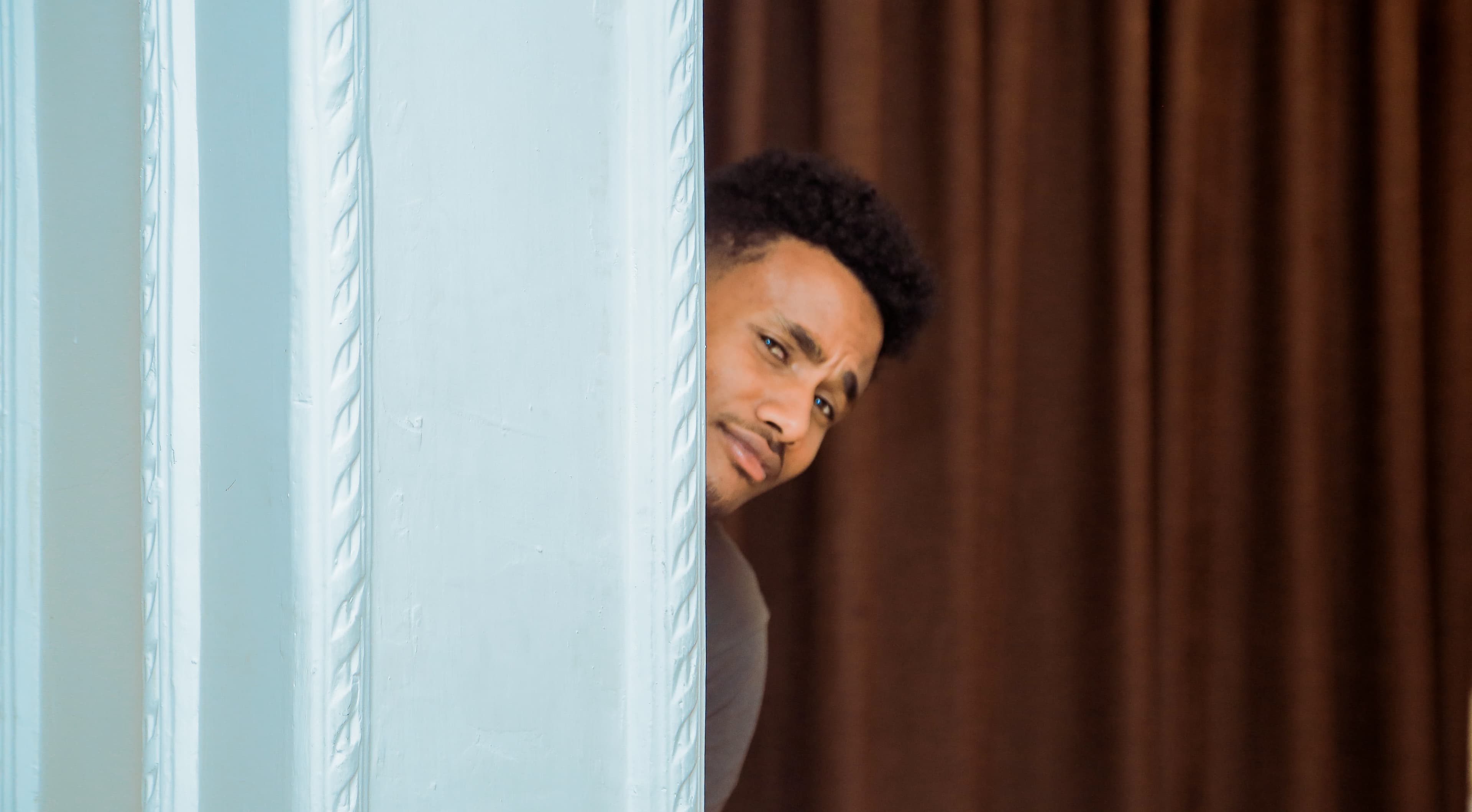 A man with curly hair and a faint mustache peeks out from behind a light blue wall, in front of a brown curtain. He is looking at the camera and has a skeptical expression on his face.