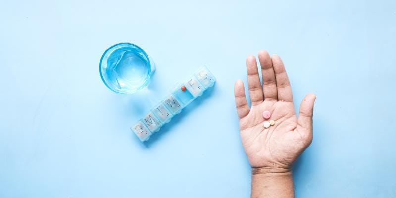 A hand holding three pills, next to a pill container and a glass of water.