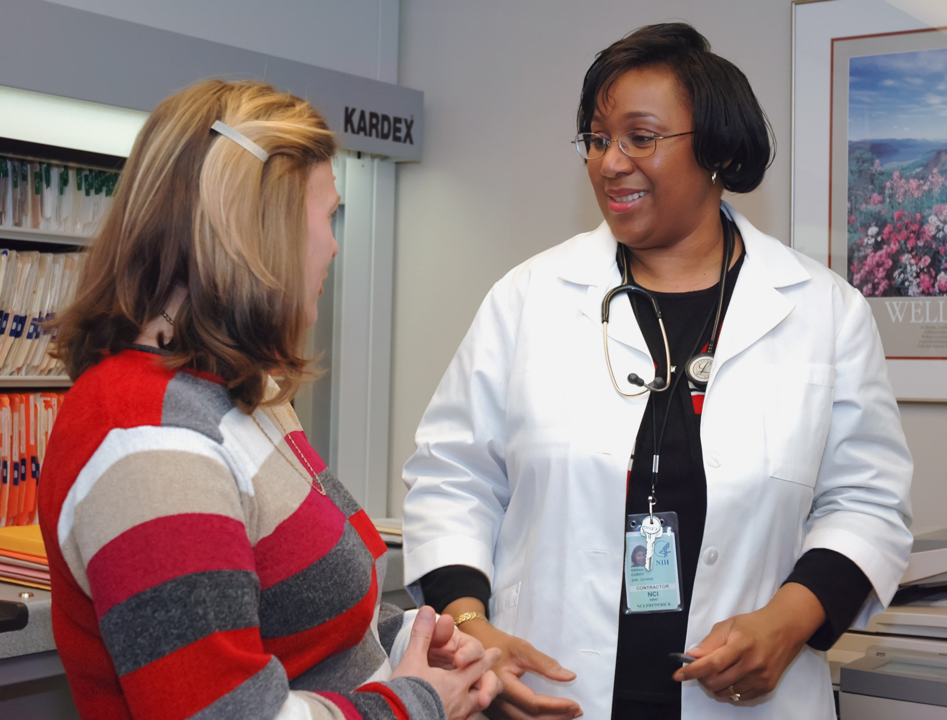 In a doctor's office, a white woman is sitting down and looking at her doctor, a smiling Black woman.