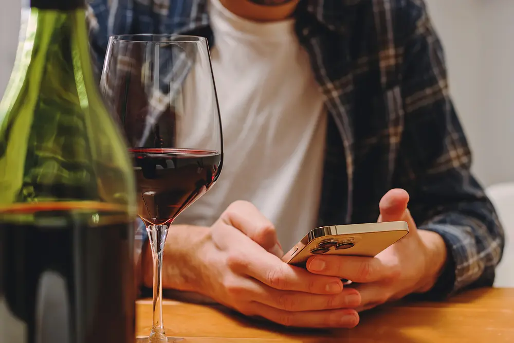 Close-up of a person’s hands texting on a phone next to a glass of wine.