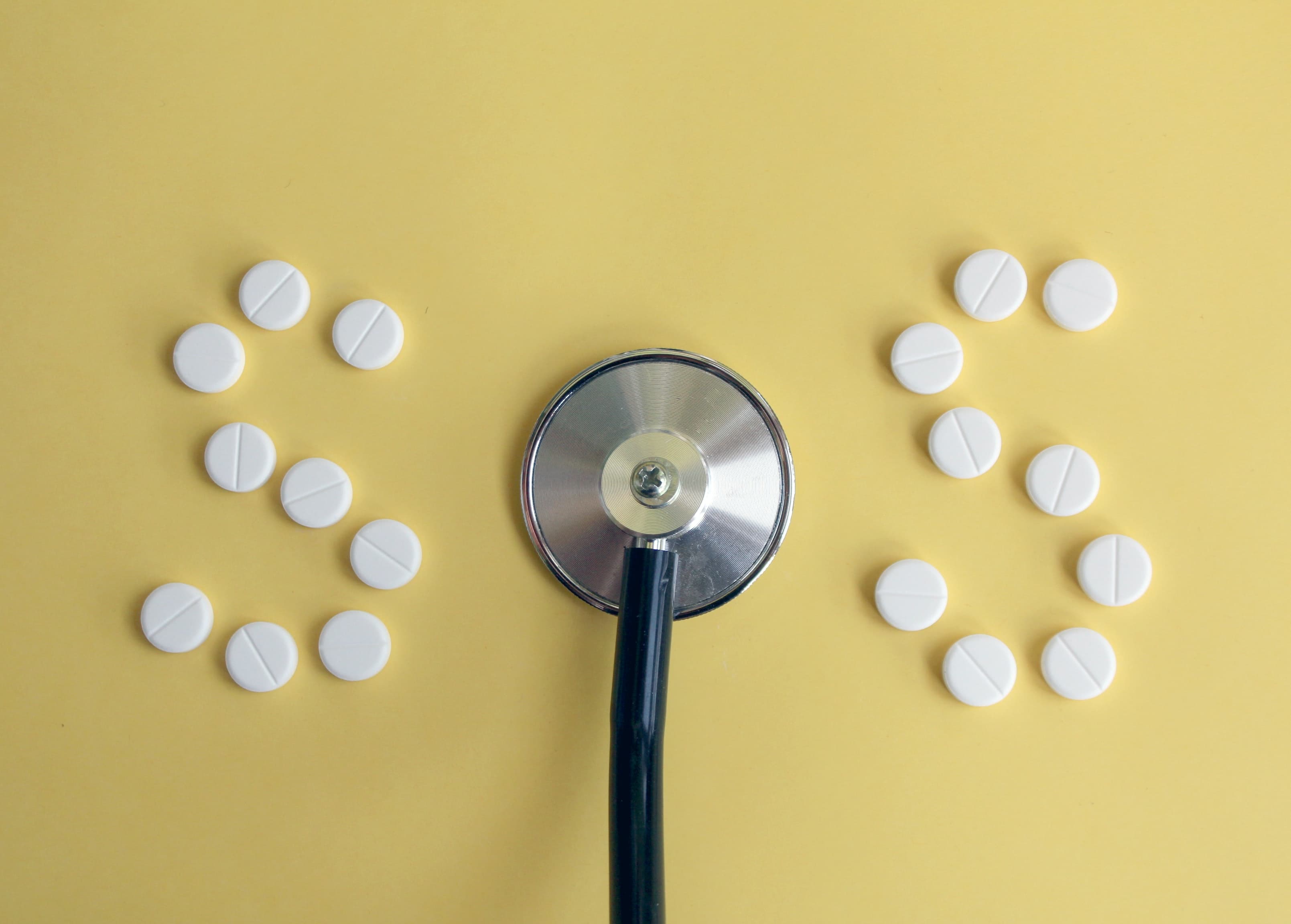 A stethoscope between two lines of white pills against a yellow background.