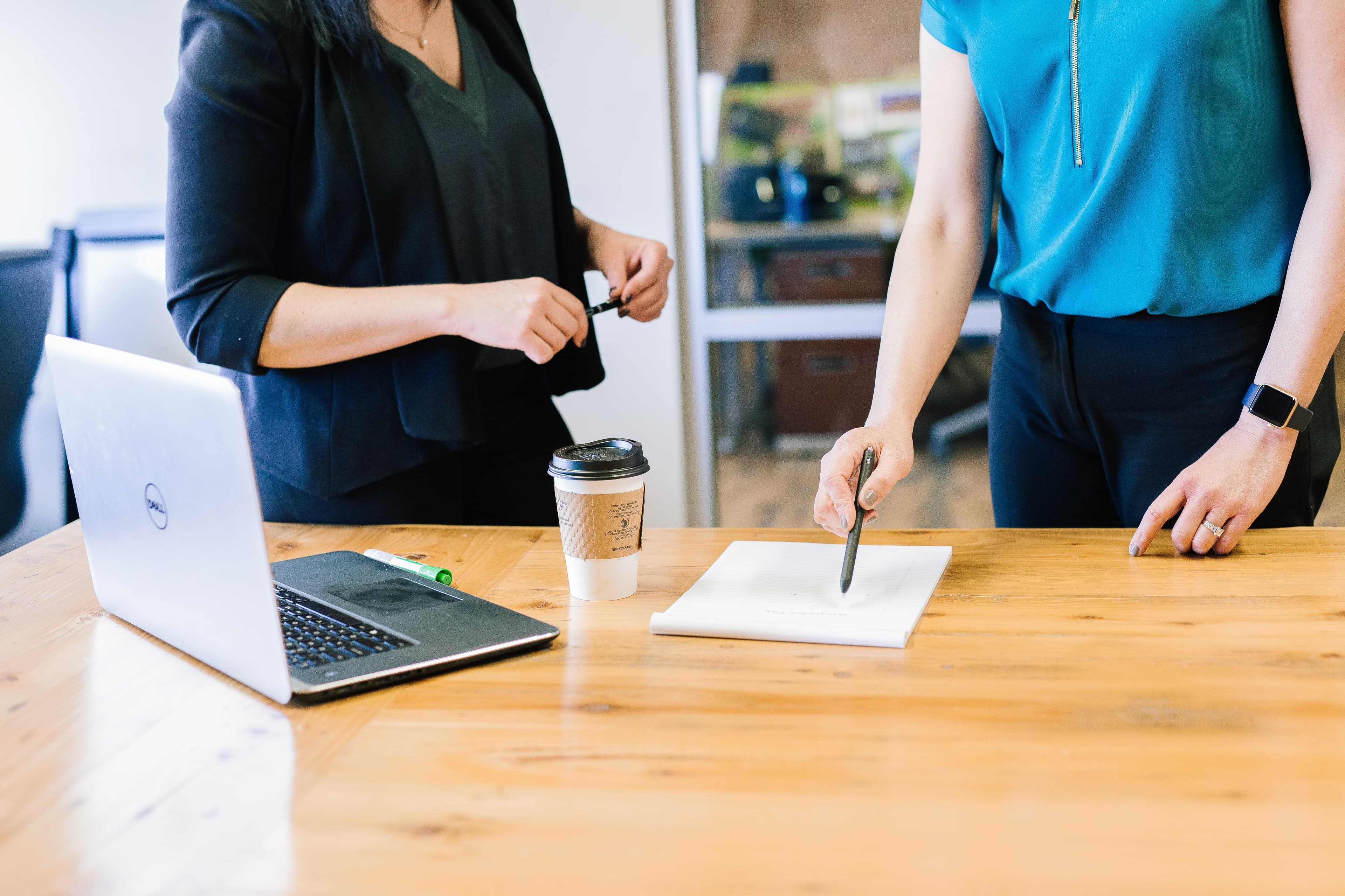 Two women stand at a conference table. They appear to be in a business meeting, and are using a laptop, papers, and pens.