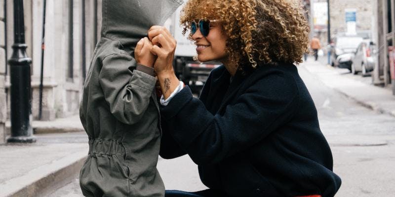 A Black woman with long curly hair wearing sunglasses, a long black coat, a red fanny pack, ripped blue jeans, and black sneakers squats on a city street. She is smiling at and holding hands with a small child. The child is wearing a gray jumpsuit and brown sneakers. The hood of the jumpsuit obscures the child's face.