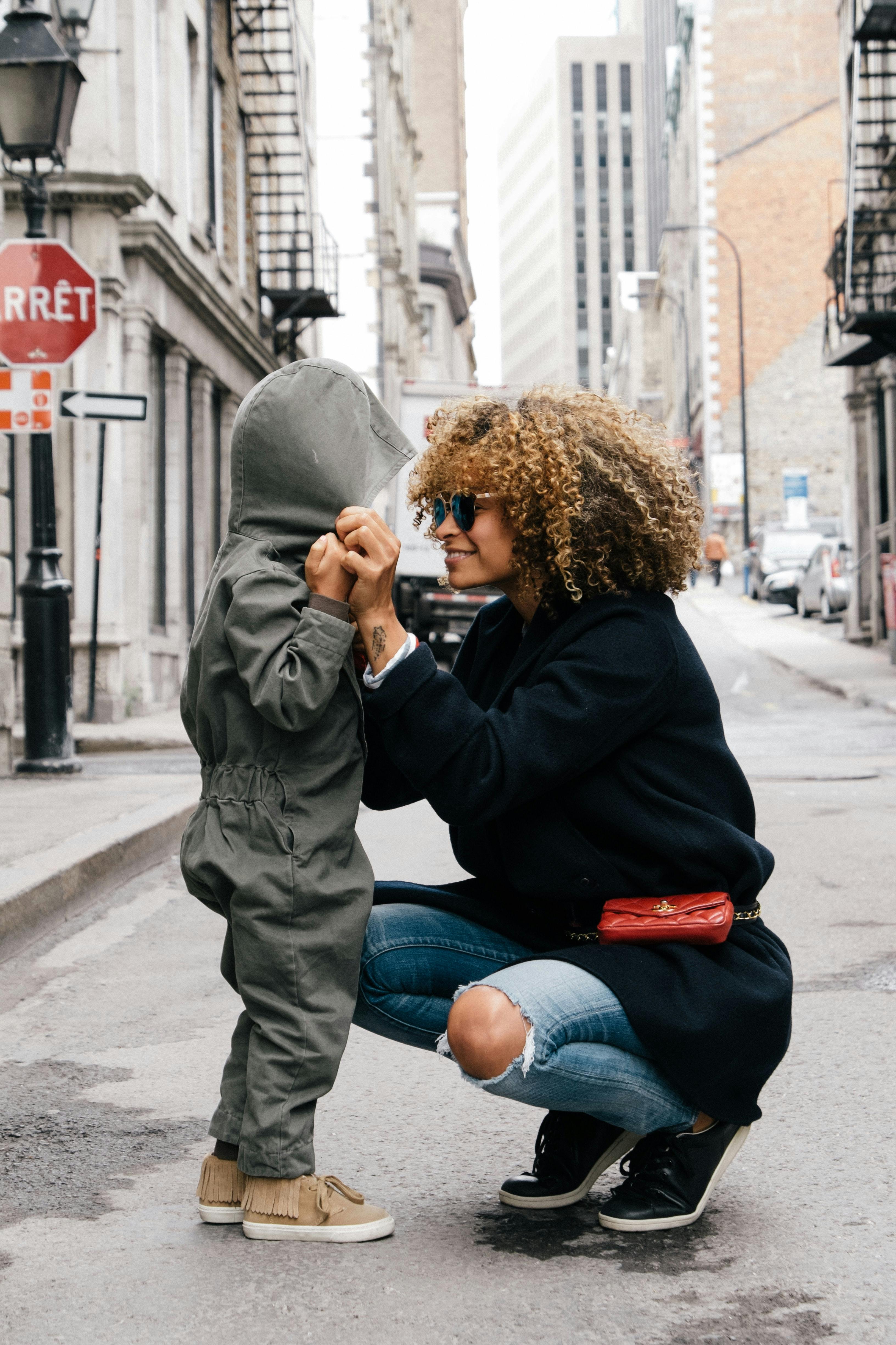 A Black woman with long curly hair wearing sunglasses, a long black coat, a red fanny pack, ripped blue jeans, and black sneakers squats on a city street. She is smiling at and holding hands with a small child. The child is wearing a gray jumpsuit and brown sneakers. The hood of the jumpsuit obscures the child's face.